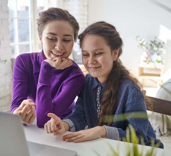 Candid shot of joyful happy young mother and daughter viewing family pictures or shopping online using laptop pc, sitting at desk in light bedroom interior, pointing fingers at screen and smiling Candid shot of joyful happy young mother and daughter viewing family pictures or shopping online using laptop pc, sitting at desk in light bedroom interior, pointing fingers at screen and smiling